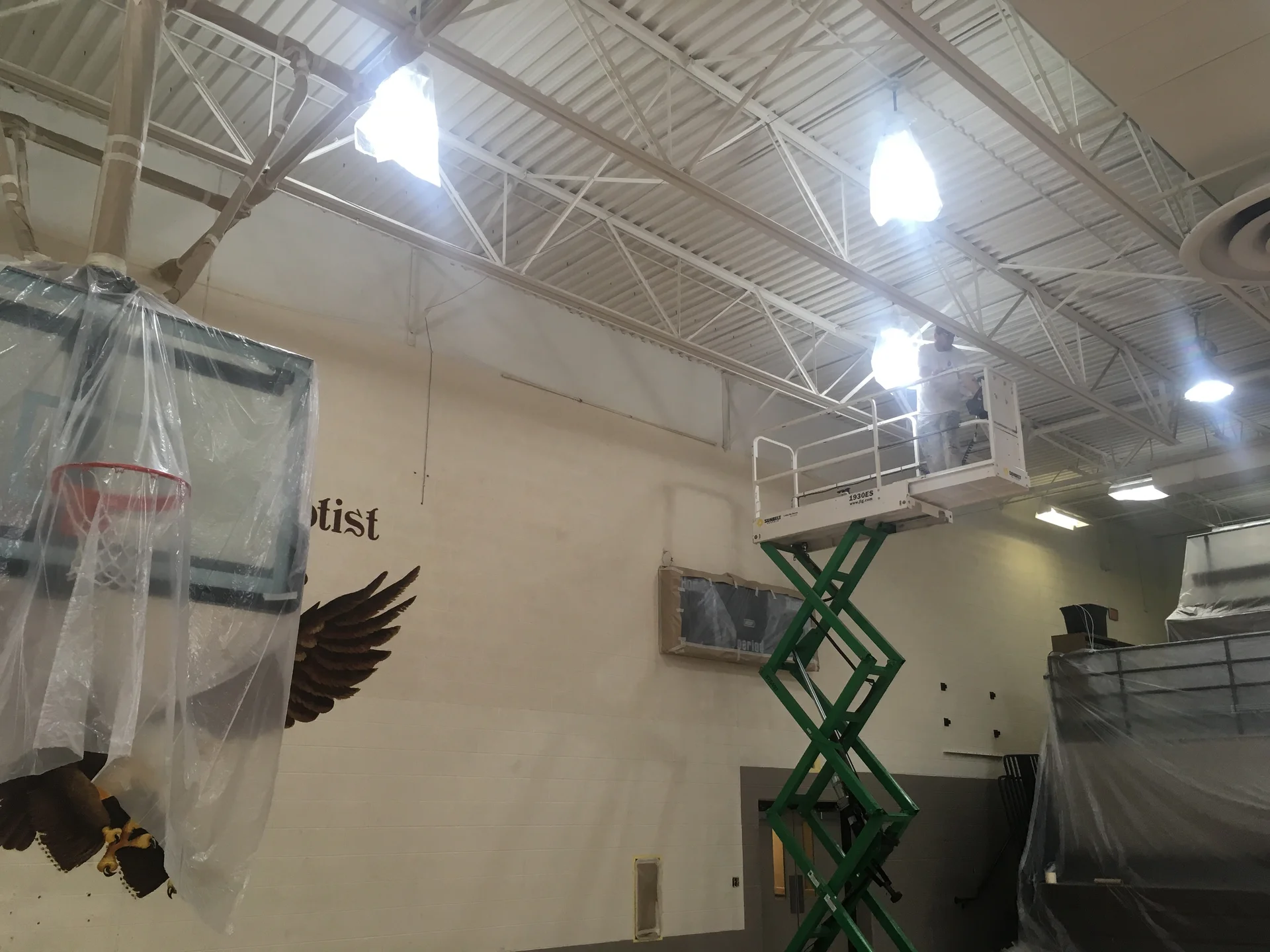 Crew on scissor lift painting upper gymnasium walls — eagle mural masked off and basketball backboard protected